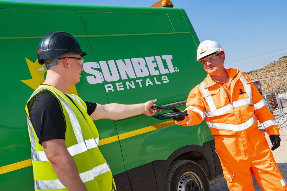 Two workers in hard hats and hi-vis passing a tablet beside a green Sunbelt Rentals van