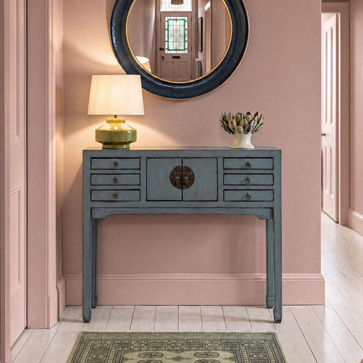 Grey-blue lacquered console table with a brass lamp and round mirror in a blush pink hallway